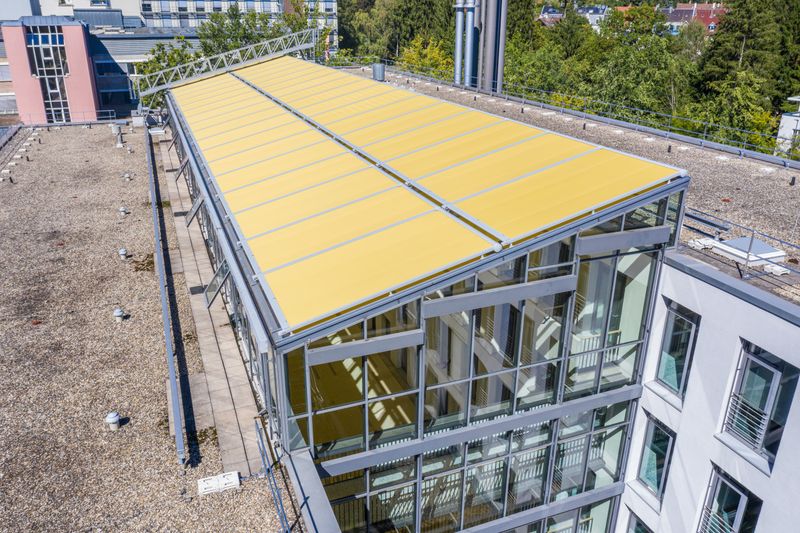 Många markiser på glas markilux 8800 med gult tyg på en korridor med glastak mellan två byggnader.