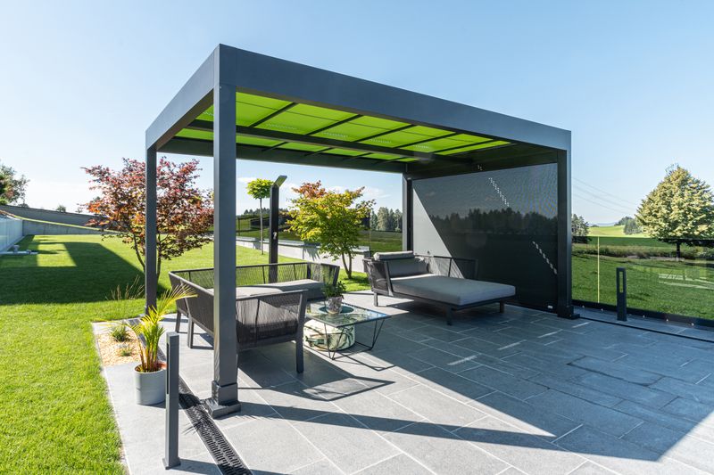 Référence d'un abri de terrasse autoportant markilux markant avec entoilage vert et store vertical avec toile translucide grise, sur une terrasse à l'écart de la maison.