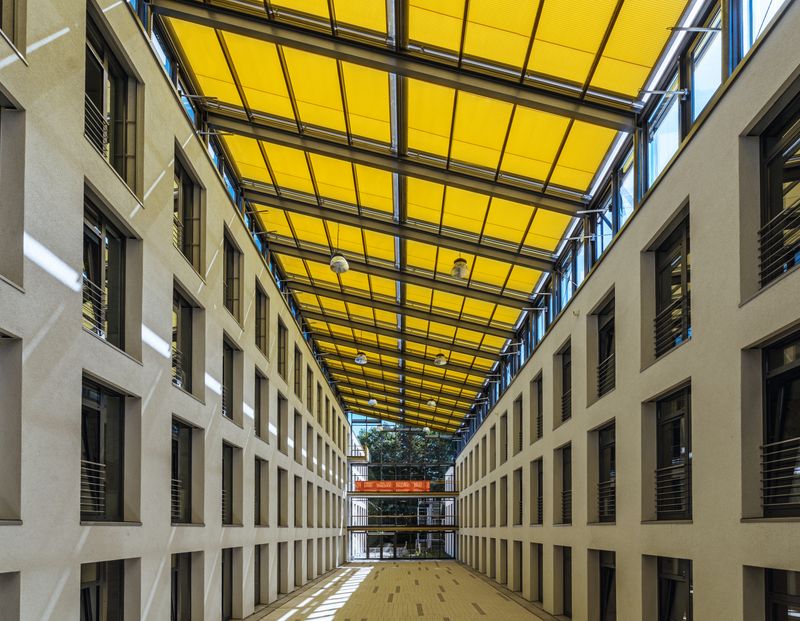Många markiser på glas markilux 8800 med gult tyg på en korridor med glastak mellan två byggnader, interiörvy.