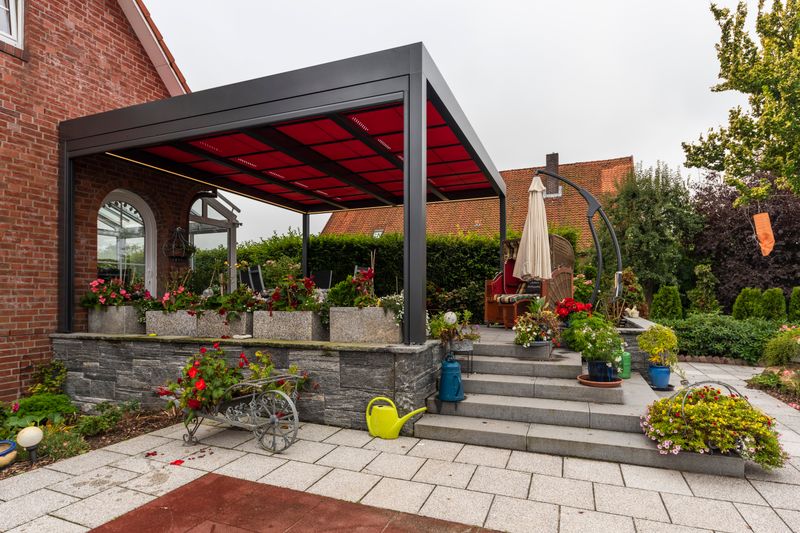 Referencia de una cubierta de patio independiente markilux markant con toldo rojo para cubrir un patio en una casa de ladrillo rojo.