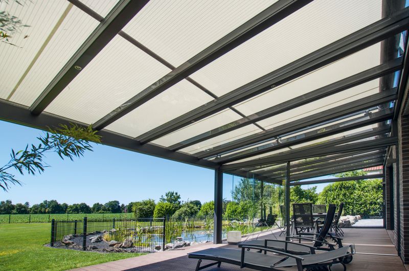 Terrace roof with on-glass awning markilux 8800, view under the roof outside to the garden with pond.
