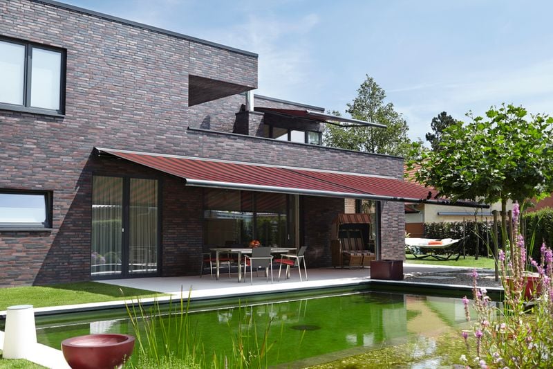 Reference image: cassette awning markilux 6000 (frame anthracite, fabric cover red-black striped) on a house with dark clinker brick, pond in the foreground.