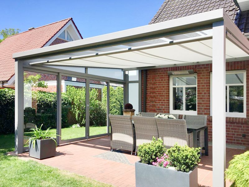 Brick house with stone terrace, covered with a light gray patio roof. An under-glass awning serves as sun protection. The frame colour matches the patio roof, the awning cover is white.