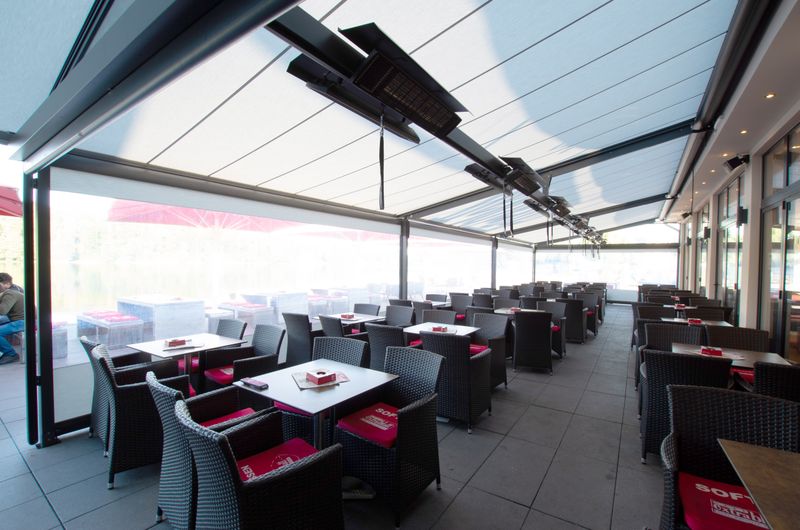 Combinação de pérgola markilux, cobertura de tecido bege e aquecedor de infravermelhos, com estore vertical com janela panorâmica para um terraço de restaurante.