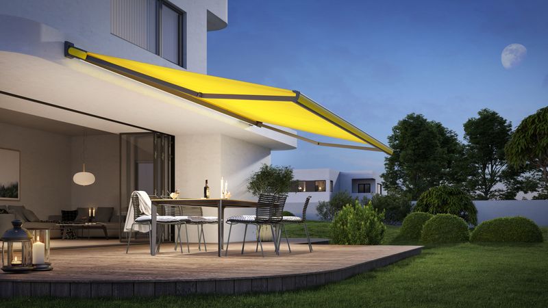 Una tenda da sole a cassonetto gialla markilux MX-3 con illuminazione LED integrata illumina di sera una terrazza in legno.