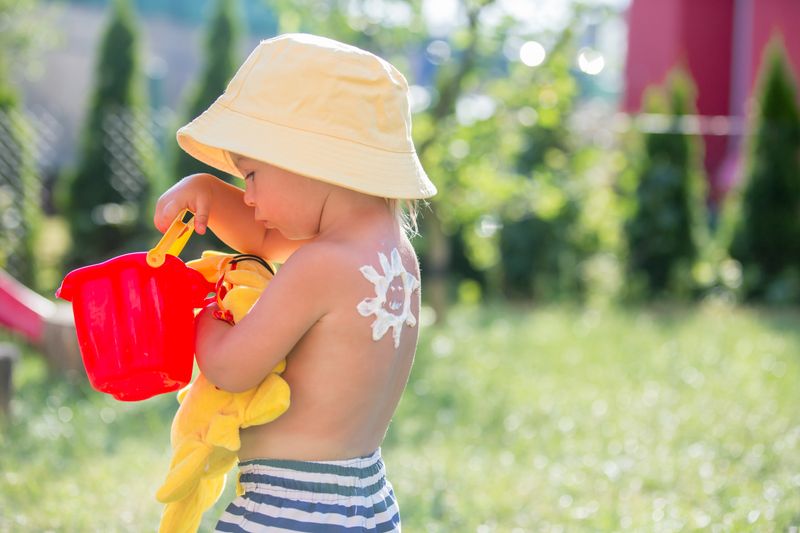 Kleinkind in Badehose und mit Sonnenmütze sowie einer Sonnencremesonne auf dem Rücken.
