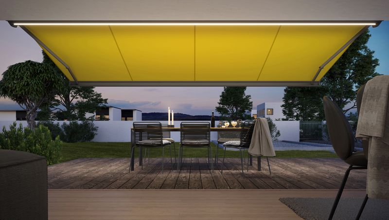 Vue de l'intérieur d'un store à coffre jaune markilux MX-3 avec éclairage LED intégré éclairant une terrasse en bois le soir.