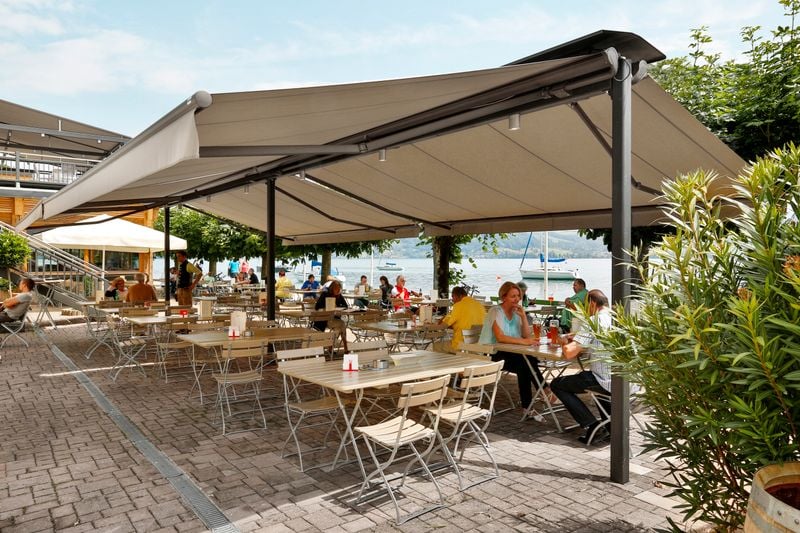 Two brown markilux 1710 with beige fabric cover and valance on a markilux syncra above the terrace of a lakeside café.