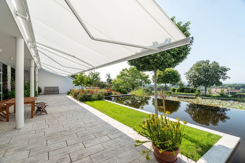 Coupled white cassette awnings markilux 6000 over a terrace adjacent to a pond.