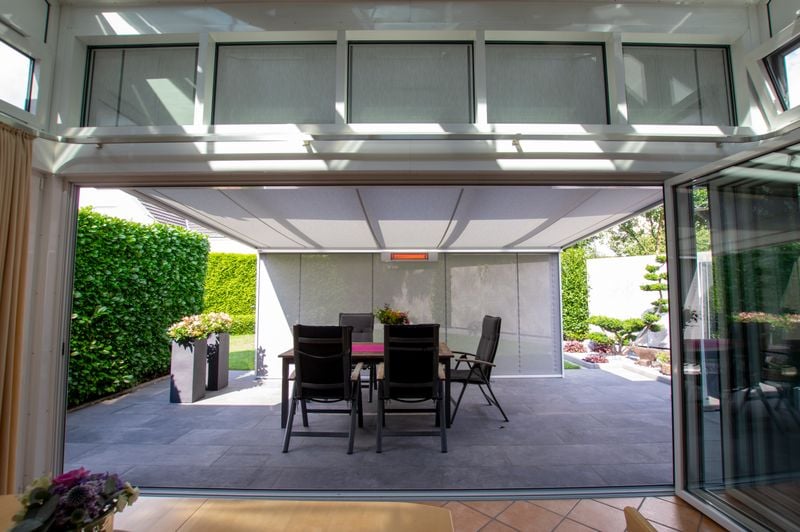 View from the living room to a terrace, which is covered with a white terrace roof with under-glass awning markilux 779 with white frame and gray fabric cover. In addition, a vertical blind with the same fabric pattern and an infrared heater are installed at the front.