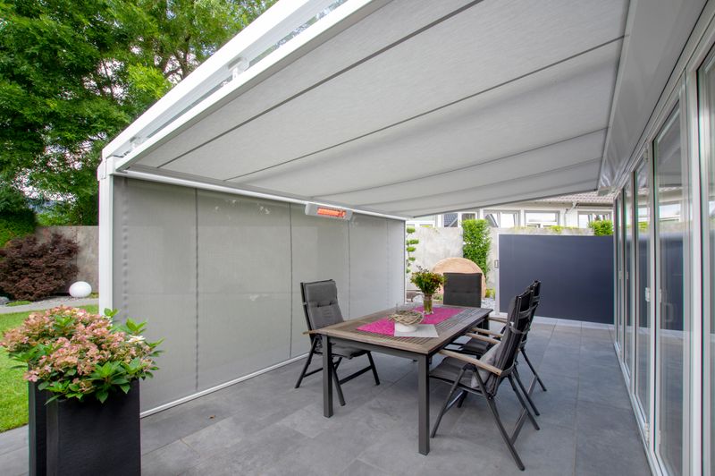 Vitt terrasstak med underglasmarkis markilux 779 med vit ram och grå tygtäckning. Dessutom är en vertikal persienn med samma tygmönster och en infraröd värmare monterade på framsidan.