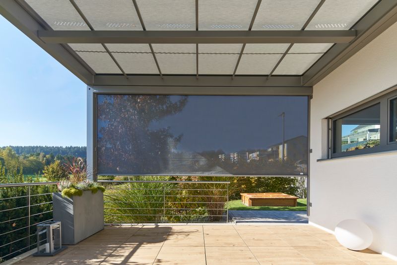 Référence d'un abri de terrasse autoportant markilux markant avec entoilage clair et store vertical avec toile translucide grise sur un bâtiment en plâtre blanc.