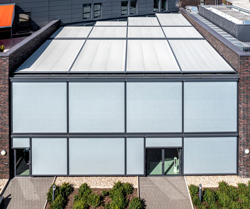 Kontorsbyggnad: mörkt tegel, platt tak, utvändig trappa, ljusgrå markilux 625 lamellgardiner som solskydd för interiören. Skuggning av hela fönsterfronten.