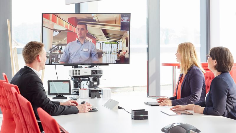 Videokonferenz dreier Mitarbeiter mit einem weiteren Mitarbeiter, dessen Bild auf einem Fernseher gezeigt wird