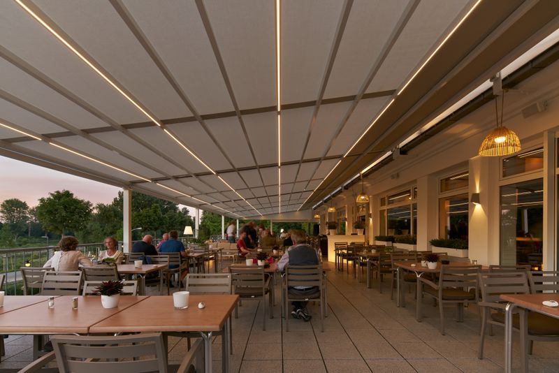 uitzicht op een restaurantterras bij schemering: markilux pergola stretch met licht doek en wit frame - voorzien van licht- en windbeschermingselementen.