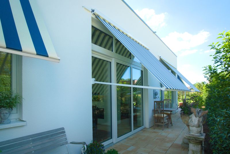 Plusieurs stores à projection markilux 730 avec toile de store rayée bleu et blanc, installés devant les fenêtres d'un bâtiment en plâtre blanc.