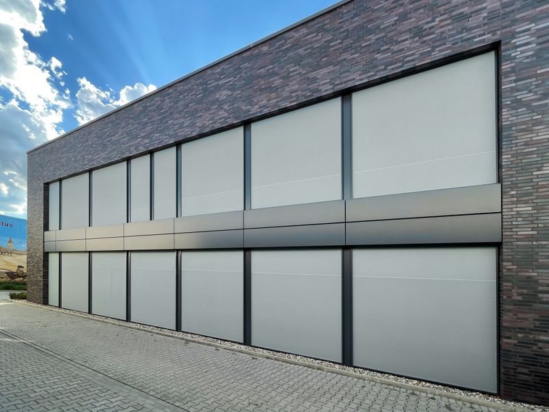 Edifício de escritórios: tijolo escuro, telhado plano, escadaria exterior, toldos verticais cinzentos claros markilux 625 como protecção solar para o interior. Sombreamento de toda a frente da janela.