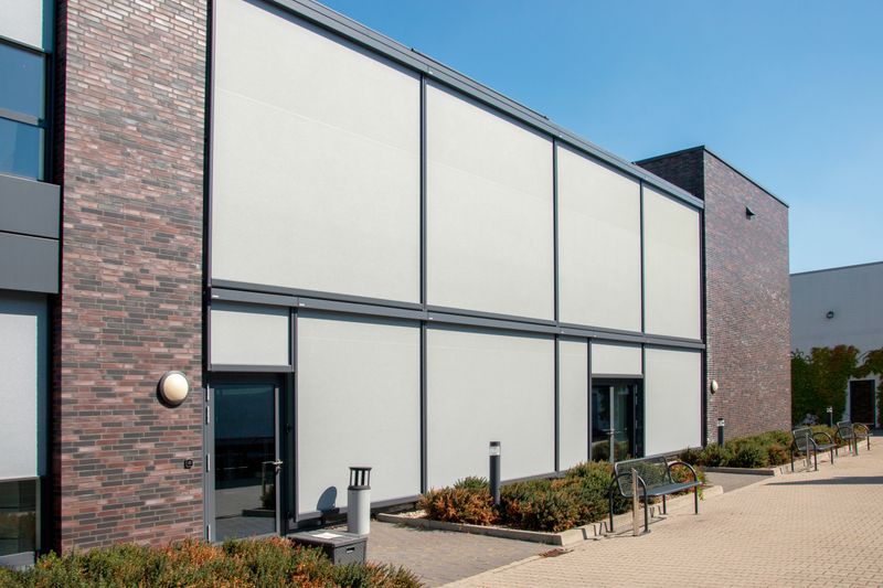 Office building: dark bricks, flat roof, external staircase, light gray markilux 625 vertical blinds as sun protection for the interior. Shading of the entire window front.