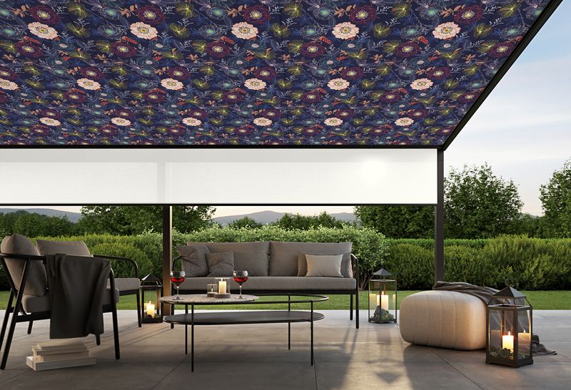 Pergola awning with dark gray frame, a fabric cover with floral pattern and white shadeplus. Cozy atmosphere with candles and two wine glasses on a table.