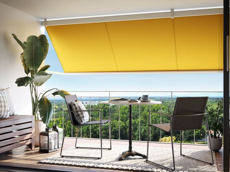 An dem Dachvorprung eines Balkons ist eine Markise mit einem gelben Tuch und einer weißen Blende angebracht.