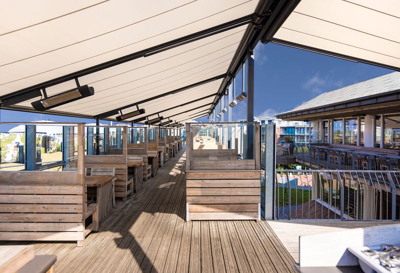 The outdoor area of a restaurant is shaded with several markilux pergola compact awnings. The frame is dark gray and the fabric covers are cream-colored. Radiant heaters are attached to the awning arms.
