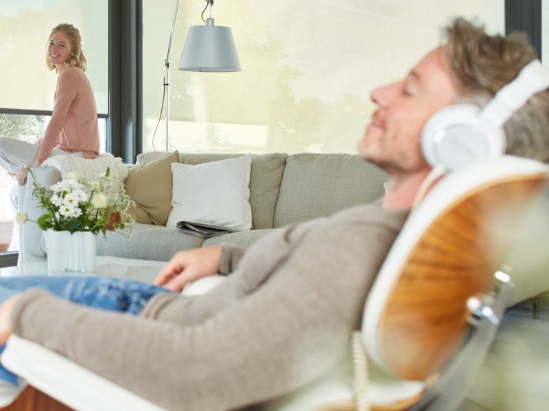 Scene i stuen: I forgrunden sidder en mand med hovedtelefoner i en lænestol, i baggrunden sidder hans kæreste på kanten af sofaen foran vinduet, som er forsynet med lamelgardiner.