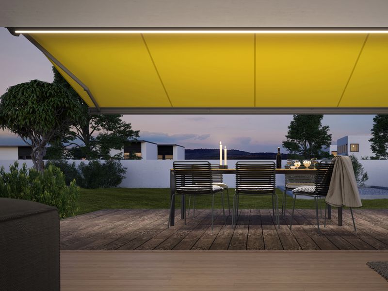 Ansicht von Innen auf eine gelbe Kassettenmarkise markilux MX-3 mit integrierter LED-Beleuchtung, die eine Holzterrasse am Abend erleuchtet.