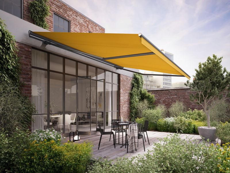 Yellow awning markilux 1710 with straight valance on a factory loft with wooden terrace and green planting.