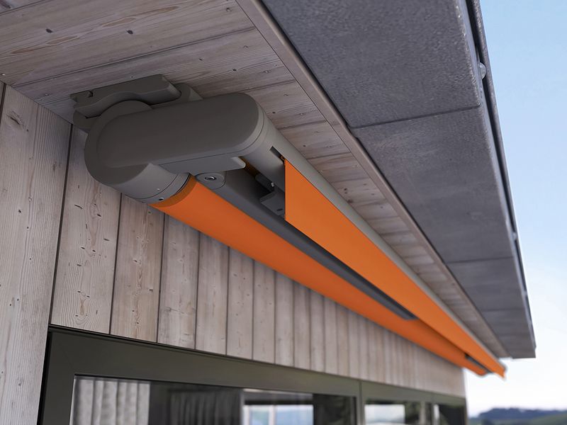 Offene Gelenkarmmarkise markilux 930 mit orangenem Tuch unter einem Dachvorstand.
