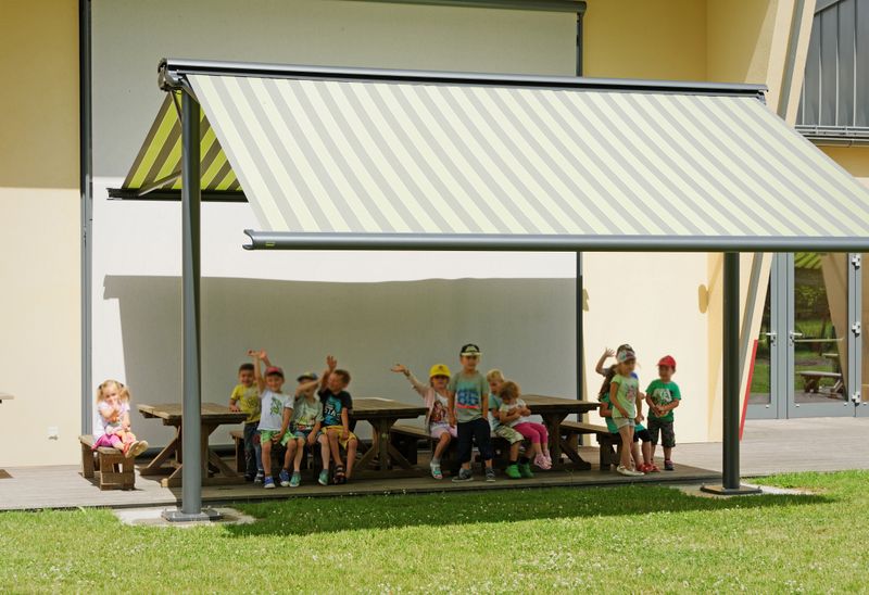 Kindergarten children sit on the terrace under a double-sided awning.