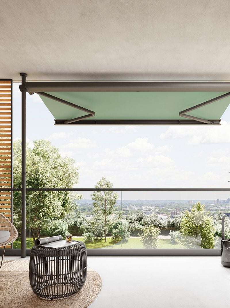 View from inside onto a modern, large balcony on which a markilux 900 clamp awning is clamped between the floor and ceiling in front of the balcony parapet. The awning with green fabric cover is extended and provides an unobstructed view of trees in the foreground and a city in the background.