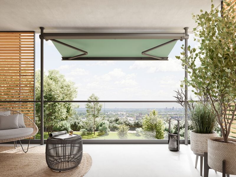 View from inside onto a modern, large balcony on which a markilux 900 clamp awning is clamped between the floor and ceiling in front of the balcony parapet. The awning with green fabric cover is extended and provides an unobstructed view of trees in the foreground and a city in the background.