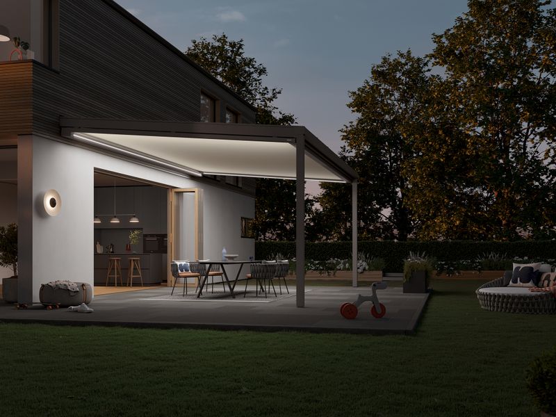 Side view of a modern detached house with a patio roof. Attached to this is the markilux 679 under-glass awning, whose integrated LED lighting illuminates the white awning cover and the patio.