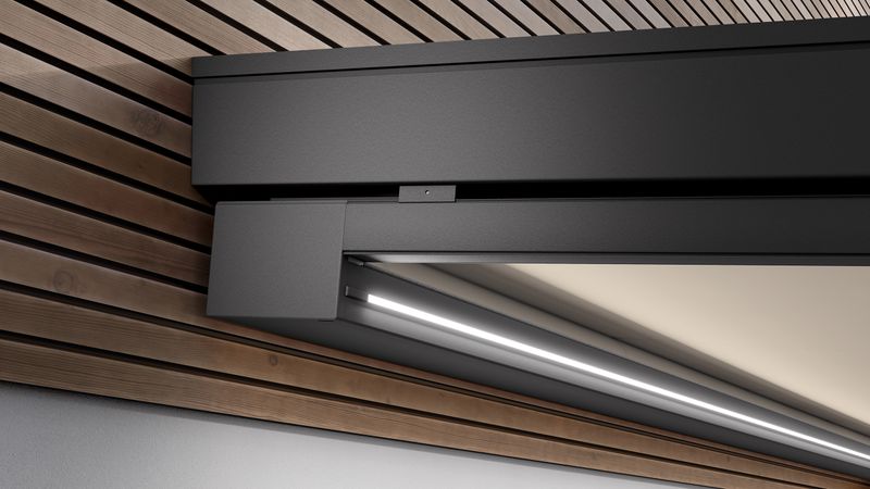 Detailed view of the cubic cassette of the markilux 679 under-glass awning with integrated LED-Line, color anthracite metallic. The under-glass awning is installed under a patio roof that adjoins a modern detached house with a wooden façade.