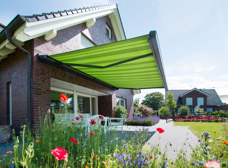 reference markilux mx-1 compact: awning with green fabric cover on a single-family house with dark clinker brick. flower meadow in the foreground.