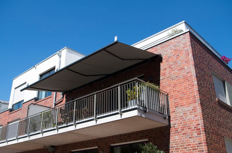 Reference image: cassette awning markilux 5010 (gray frame, gray fabric cover) covers a balcony of a modern brick house.