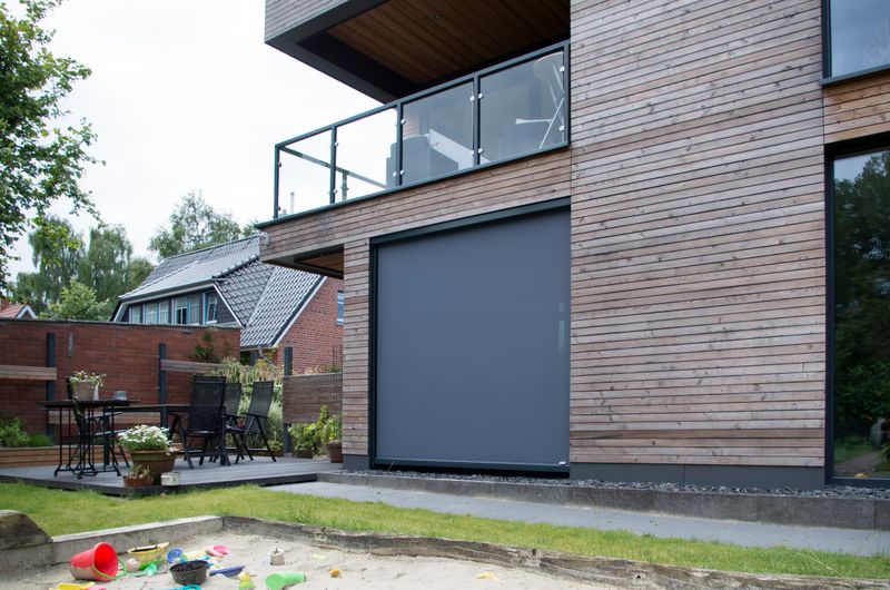 Enfamiljshus med träfasad och grå markilux-fönstermarkiser med vertikala blinderingar.