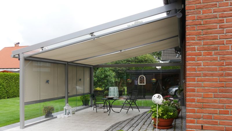 Gray patio roof equipped with a color-coordinated markilux 879 under-glass awning with light fabric cover. In front of the patio roof is an additional vertical blind 876 in the same color concept.