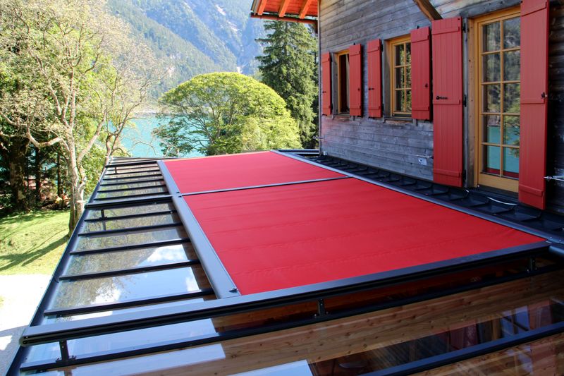 Image de référence haut store en verre markilux 8800 avec toile de store rouge, vue de dessus du tissu. Le design du store est assorti à la maison en bois aux volets rouges.