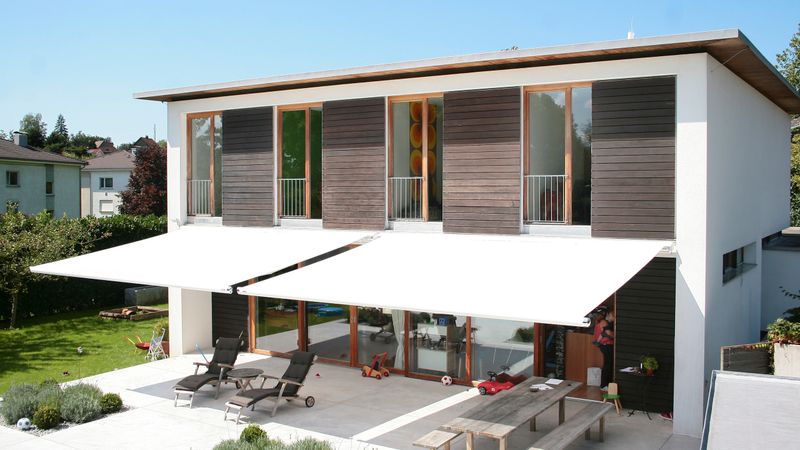 Two markilux 1600 awnings with white fabric cover over the terrace on a brown wooden façade.
