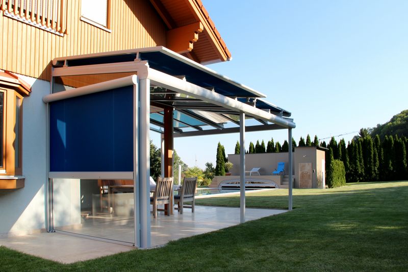 Reference image of a markilux 870 rooftop awning with gray frame and blue fabric cover on a patio roof. Side view of the terrace protected by a vertical blind on the side.
