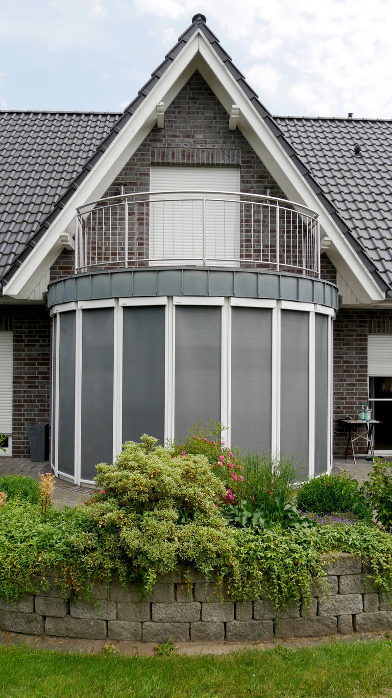 Vertical blaind awning markilux 620 with white frame and gray fabric cover attached to a round conservatory of a family house.