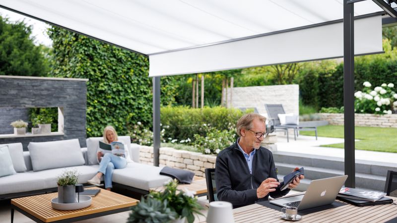 Terras met glazen dak en wit onderglasscherm. Vrouw zit in een lounge en leest. Man werkt aan een tafel.