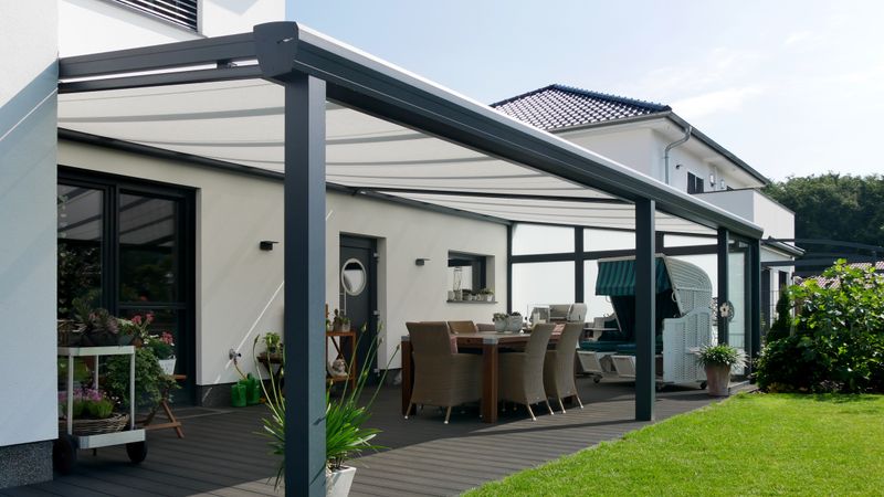 White rendered house with wooden terrace and anthracite-colored awning with markilux 879 under-glass awning. The frame of the awning matches the color of the terrace roof, the awning cover picks up the white color scheme of the house.