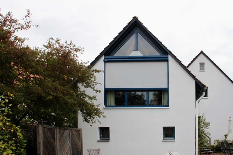 Maison blanche avec fenêtre à pignon triangulaire, store de fenêtre à demi déployé markilux 893, triangulaire.