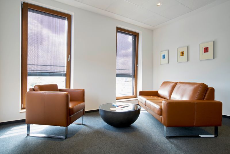 Objeto de referencia: zona de espera con sofá y sillón de cuero marrón en un edificio de oficinas. La zona de asientos está muy cerca de las ventanas, que tienen un toldo vertical markilux 620 con lona de tejido translúcido gris.