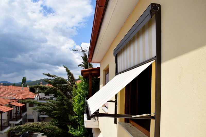 Image de référence : store mi-vertical mi-projection 740 avec toile de tissu rayé blanc et jaune et cadre noir, fixé devant la fenêtre d'une maison jaune.