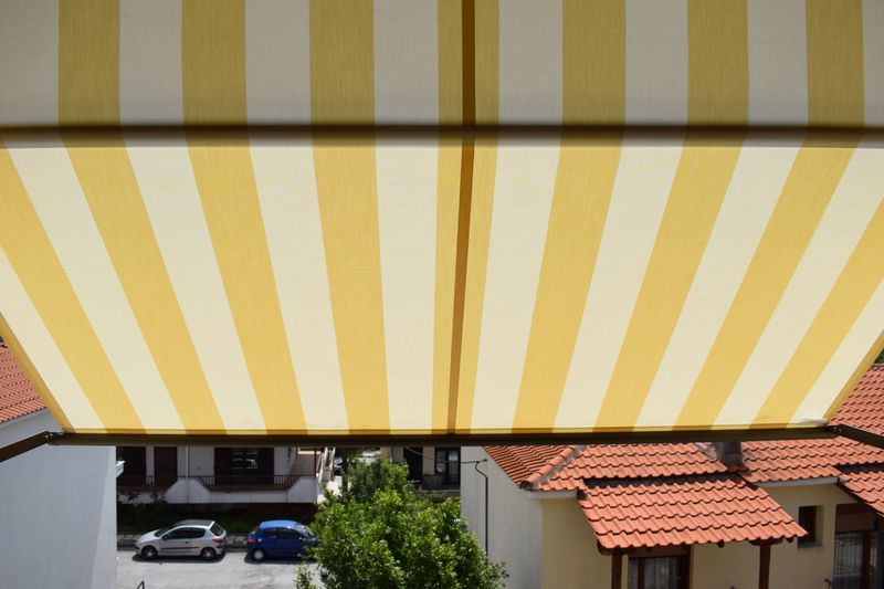 Image de référence : Vue intérieure d'une marquisolette markilux 740 avec toile de tissu à rayures blanches et jaunes, vue de la rue en contrebas de la marquisolette.