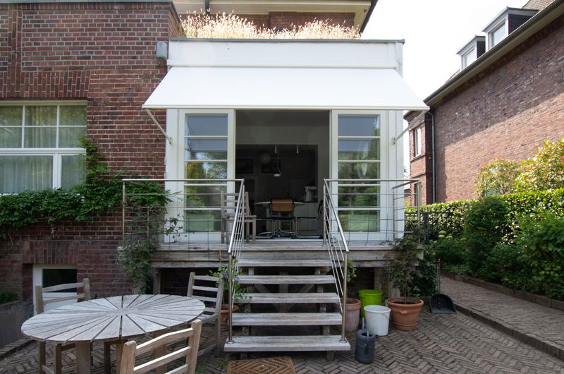 White drop-arm cassette awning markilux 730 fixed in front of a window, above a raised terrace connected by steps to the garden.