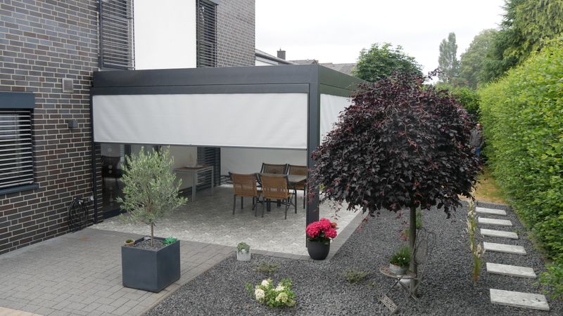 Referens av ett fristående terrasstak markilux markant och vertikal persienn med lätt markisskydd för skuggning av en terrass på ett grått tegelhus.